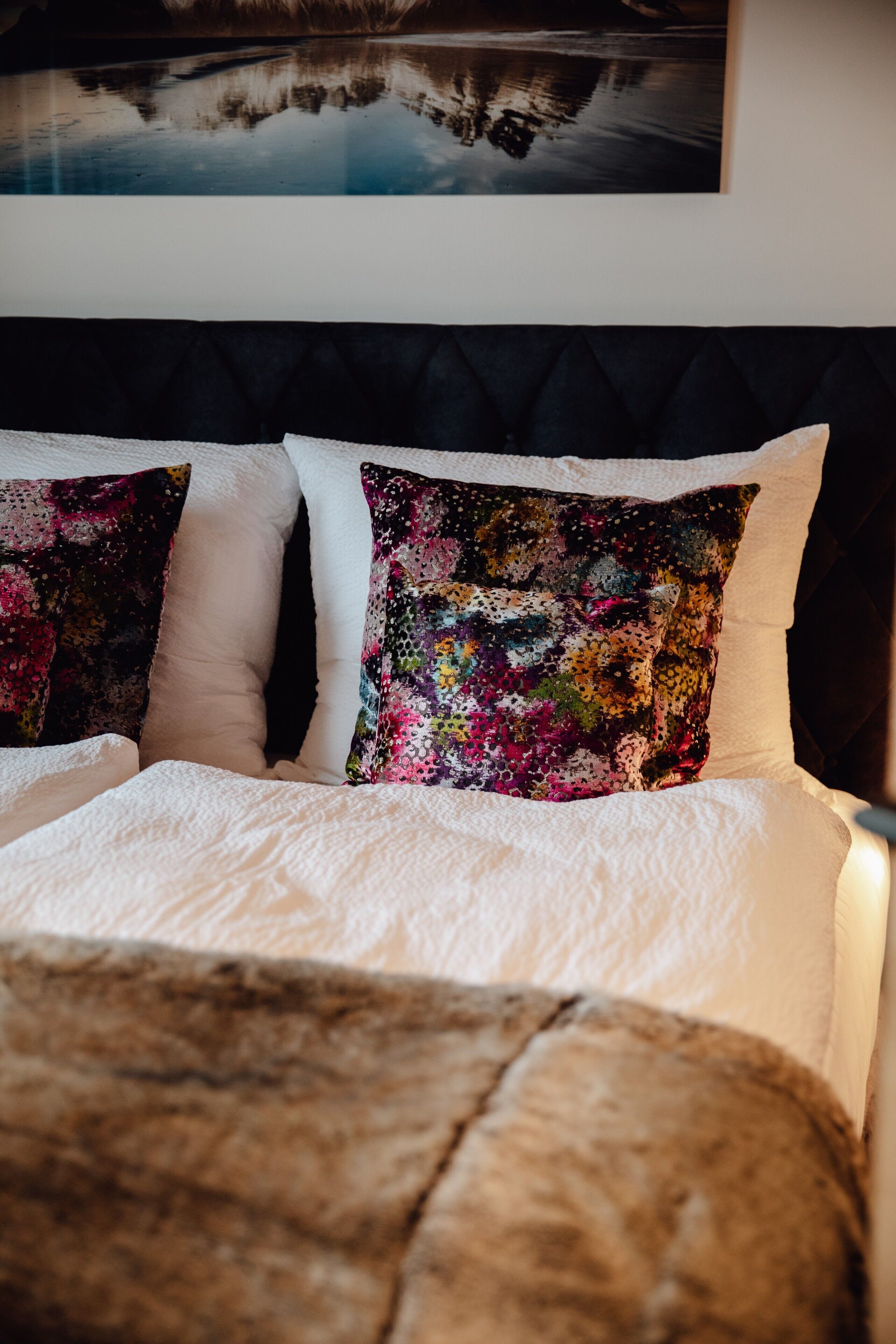 A bed with white linens and two decorative pillows, a dark headboard, and a framed picture above.