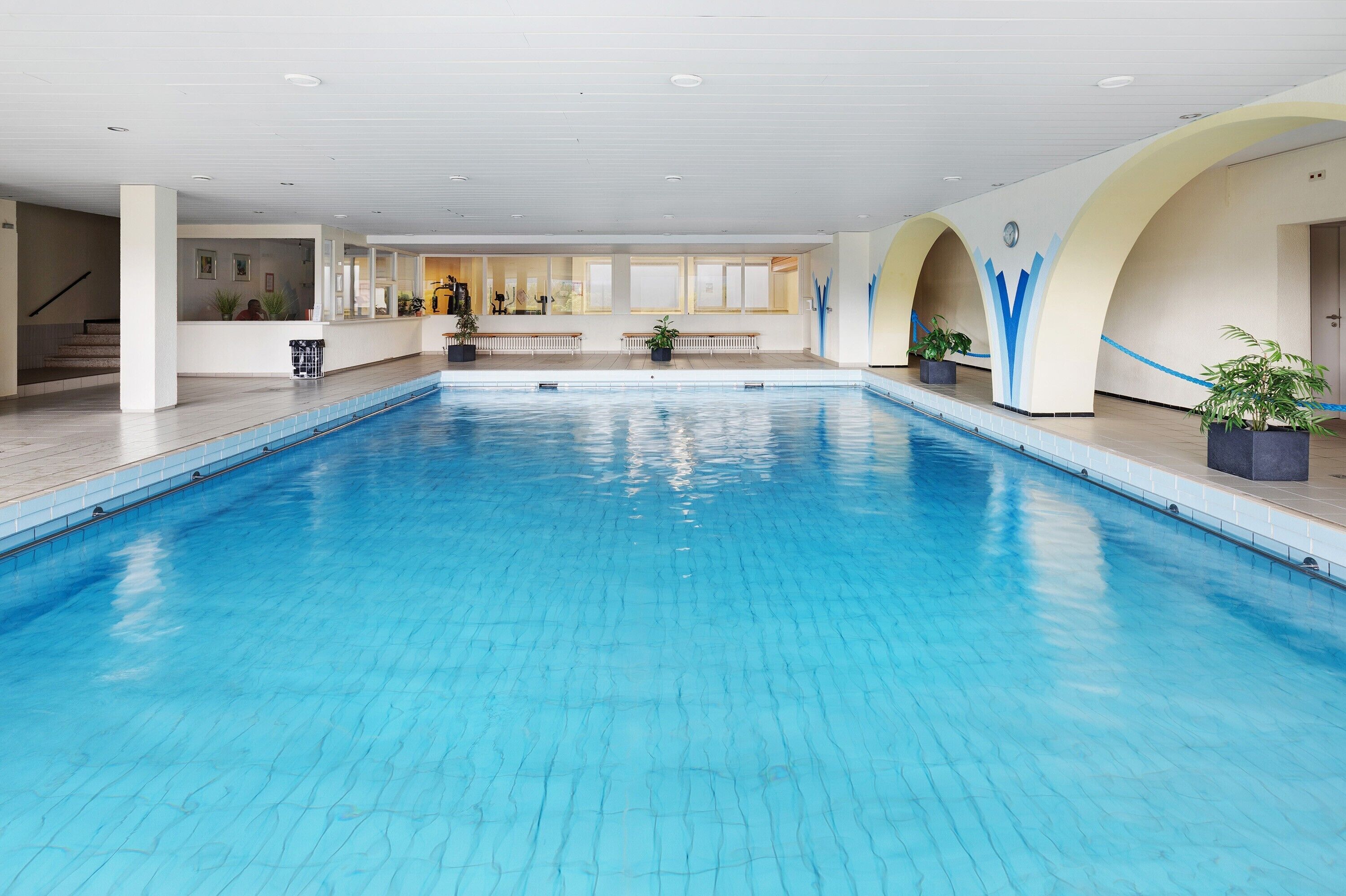 An indoor swimming pool with clear blue water, surrounded by white walls with blue accents, and a seating area with benches.