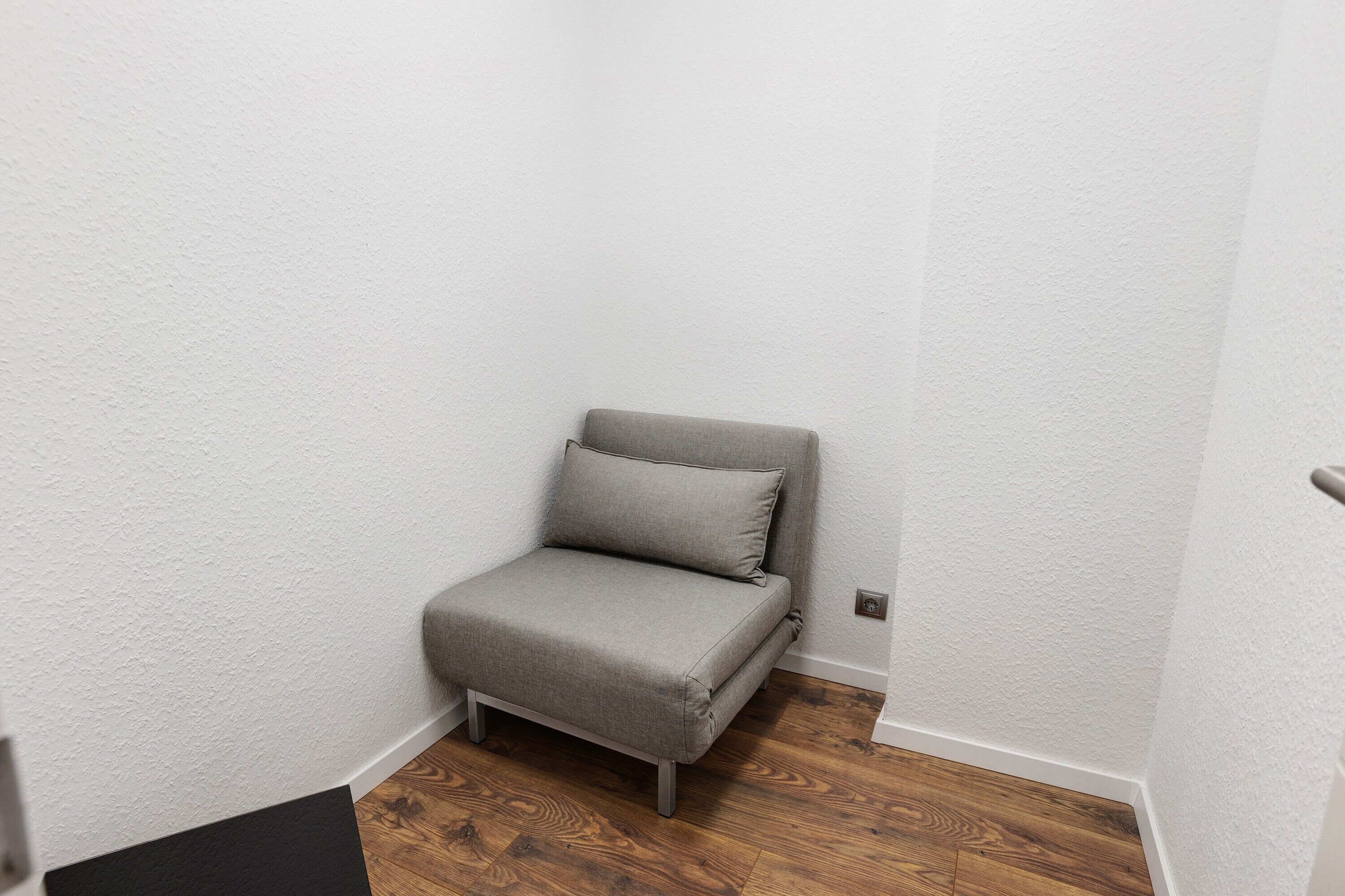 A small, narrow room with a grey upholstered chair against a white wall, wooden flooring, and a door on the right.