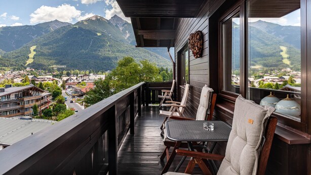 Ein Balkon mit Holzwerteln, einem Tisch und zwei Stühlen mit Blick auf eine Berglandschaft und eine darunter liegende Stadt.
