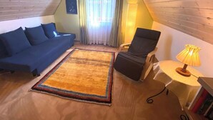 Une pièce mansardée accueillante, avec un canapé bleu, un fauteuil noir, une petite table avec une lampe et un tapis coloré.
