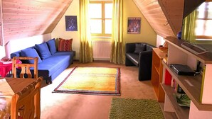 Une chambre à coucher confortable, aménagée en mezzanine, avec un plafond en pente, un canapé bleu, une chaise en bois, une petite table, un téléviseur et une bibliothèque.