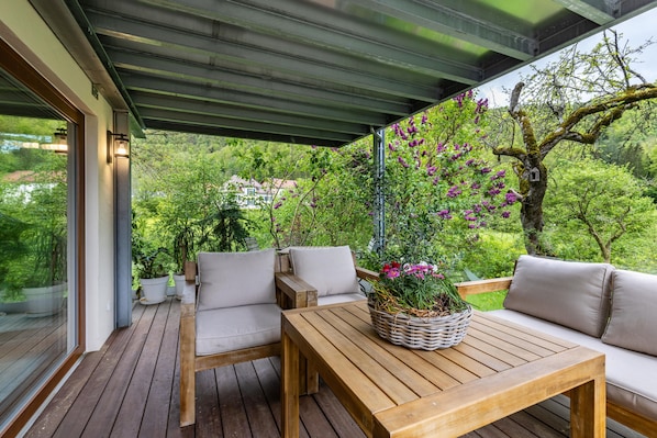 An outdoor deck with wooden furniture, a table with a basket of flowers, and a view of greenery and trees.
