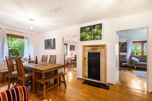 A dining area with a wooden table and chairs, a fireplace, and a view of greenery outside.