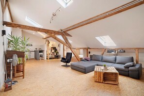 Une chambre mansardée avec un plafond en pente, des poutres apparentes, un canapé d’angle gris, une table basse en bois et une plante en pot.