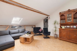 Ein Loft-Wohnzimmer mit schrägem Dach, einem Ecksofa, einem kleinen Holztisch, einem schwarzen Sessel, einer Vitrine mit Büchern und einer Topfpflanze.