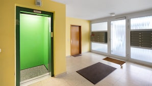 Eine Hotelhalle mit einem grünen Aufzug, einer Holztür, einem Empfangstresen und einem großen Fenster mit Jalousien.