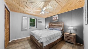 A bedroom with a wooden ceiling, a bed with a white and black patterned bedspread, a nightstand with a lamp, and a window with blinds. - Cozy Muskoka cottage (Bracebridge)