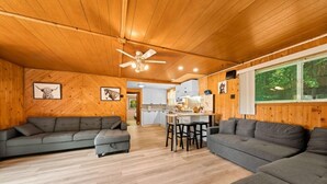 A cozy cabin interior with a wooden ceiling, a gray sectional sofa, a dining area, and a kitchen in the background. - Cozy Muskoka cottage (Bracebridge)