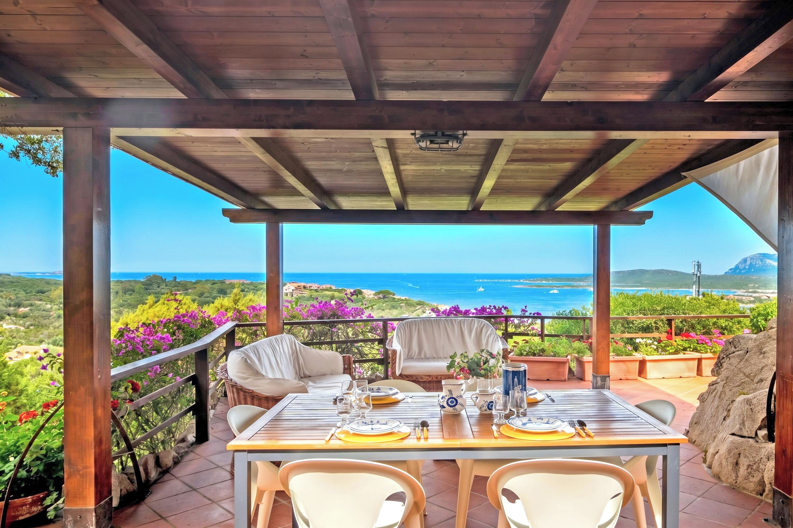 Un área de comedor al aire libre techada, con una mesa preparada para dos, con vistas panorámicas al mar y las colinas.