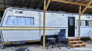Una caravana antigua estacionada bajo un refugio de madera con una parrilla y una pequeña mesa.