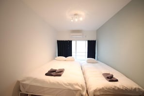 Una habitación pequeña y limpia con dos camas, una ventana con cortinas, un aire acondicionado y una lámpara en el techo.