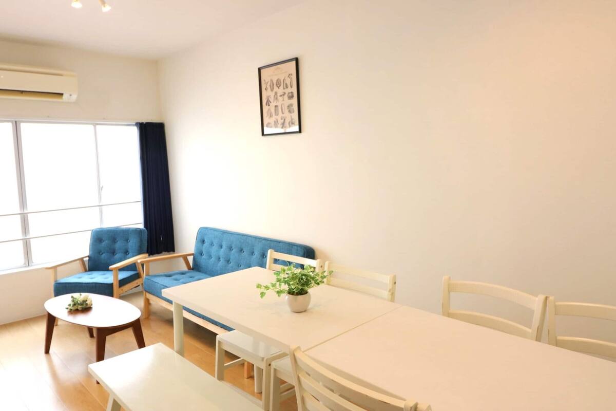 A room with a dining table, chairs, a blue sofa, a small round table with a plant, and a window with curtains.