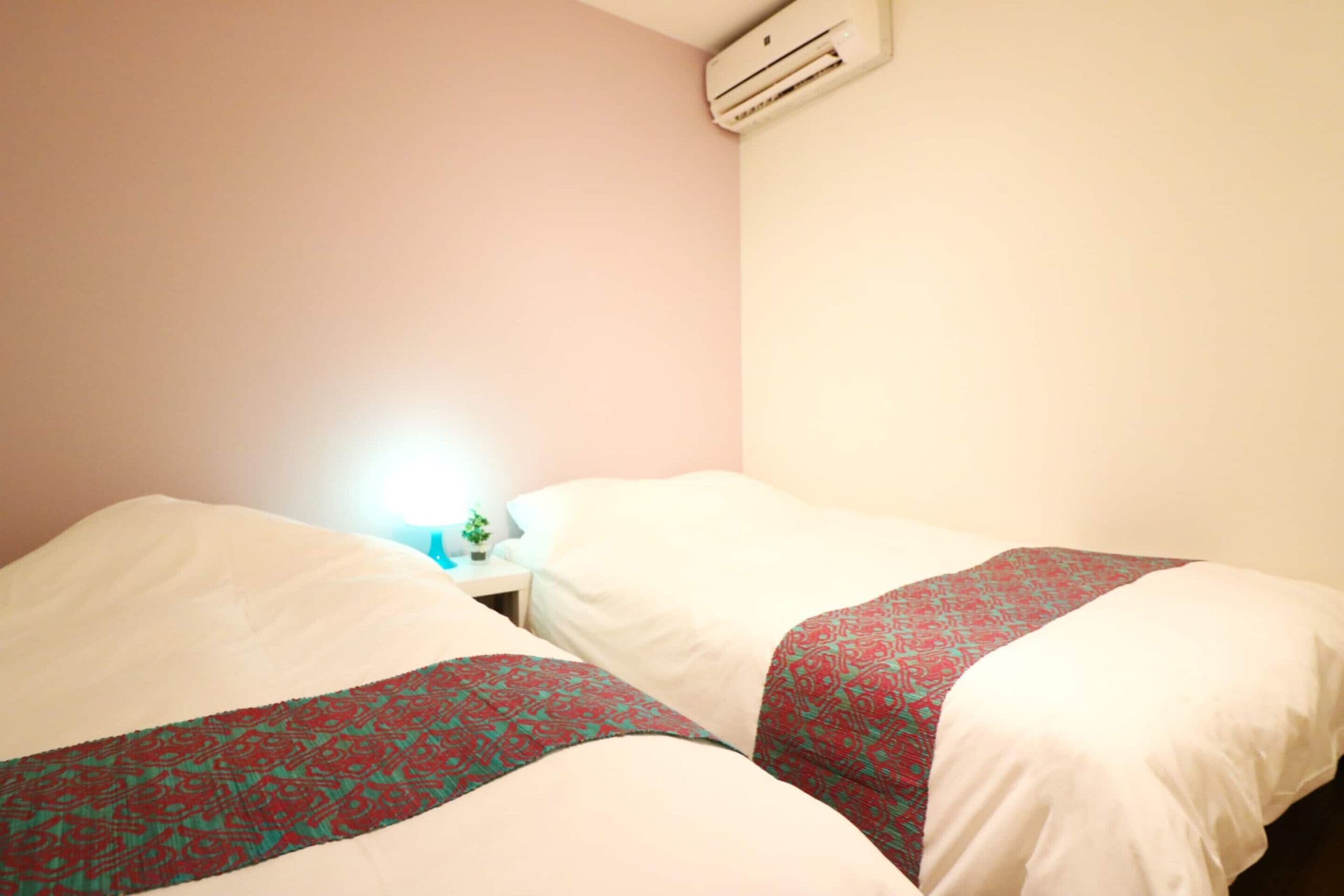 Une chambre avec deux lits, une table de chevet avec une lampe et un appareil de climatisation fixé au mur.