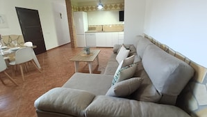 A living room with a sofa, a dining table with chairs, a kitchen area, and a door.