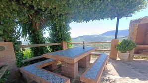 An outdoor patio with a brick table and benches, surrounded by greenery and a view of distant hills.