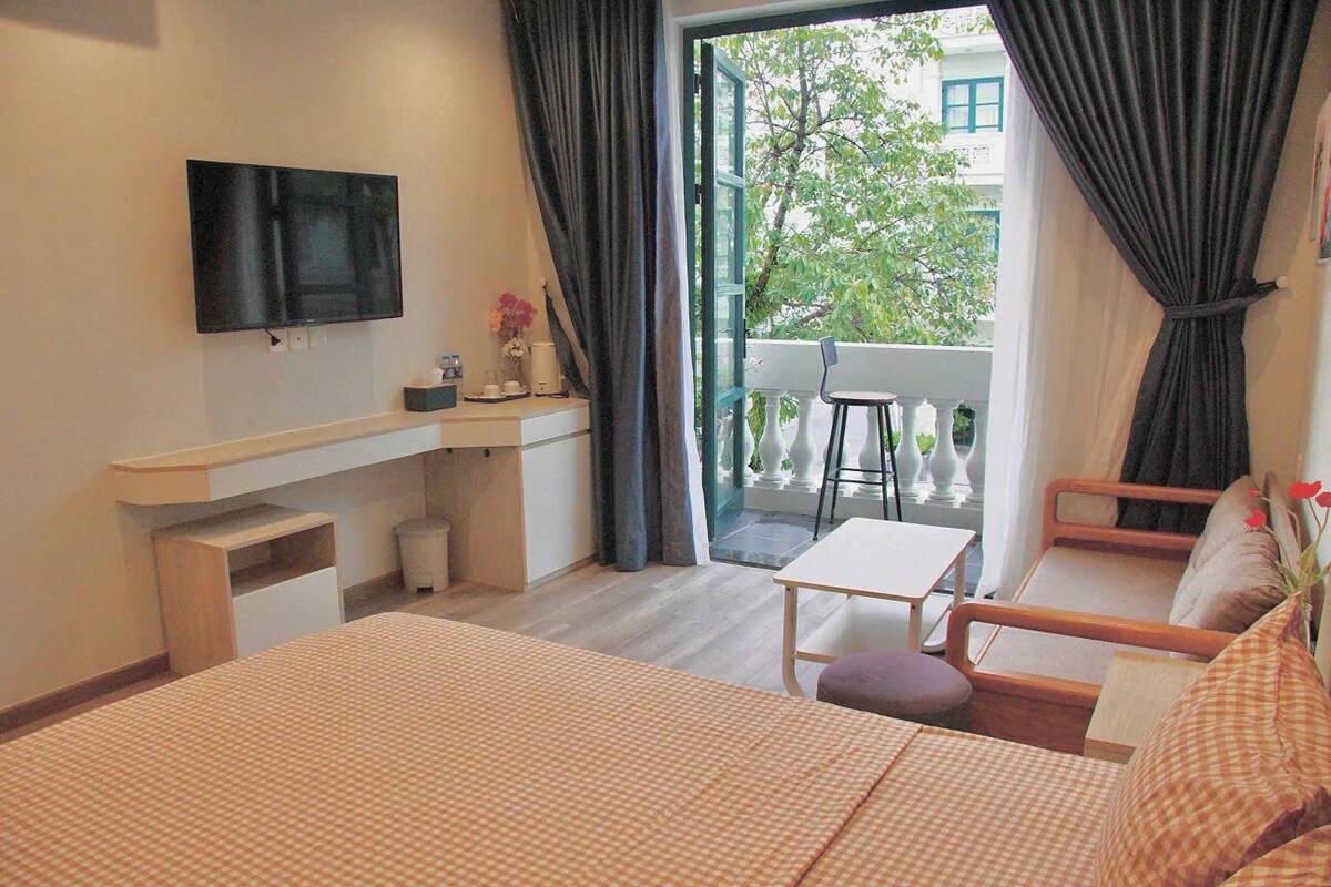 A hotel room with a bed, a desk, a TV, a balcony with a chair, and a view of the outside.