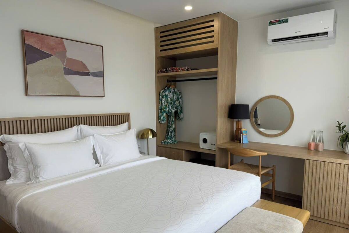 A bedroom with a bed, a desk, a mirror, an air conditioner, and a wall-mounted artwork.