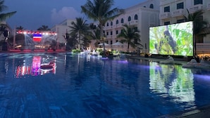 A swimming pool with a large screen displaying an image, surrounded by palm trees and a building with balconies. - Minh Anh Hotel (Phu Quoc)