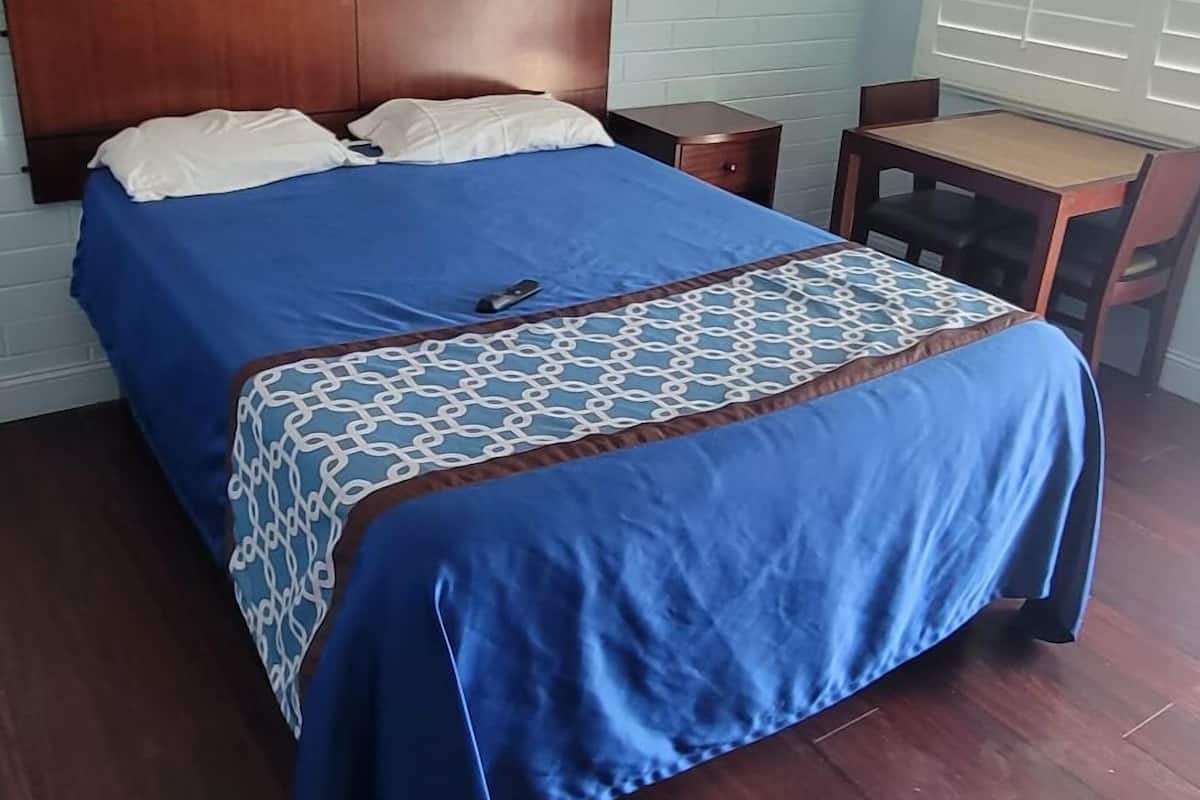A bedroom with a wooden headboard, a bed with blue bedding, a bedside table, and a small wooden desk with a chair.