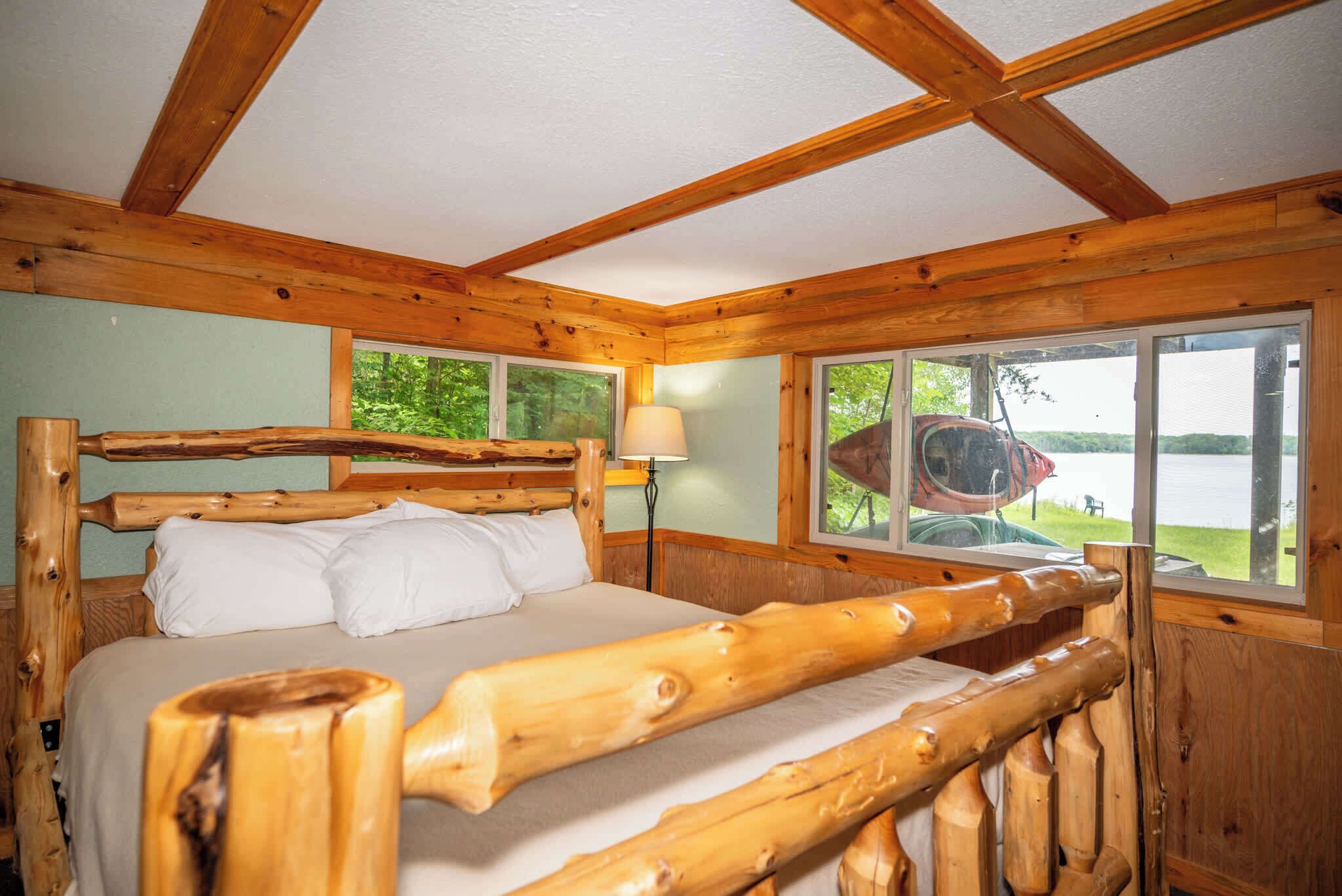A wooden cabin bedroom with a bed, windows, and a view of a lake.