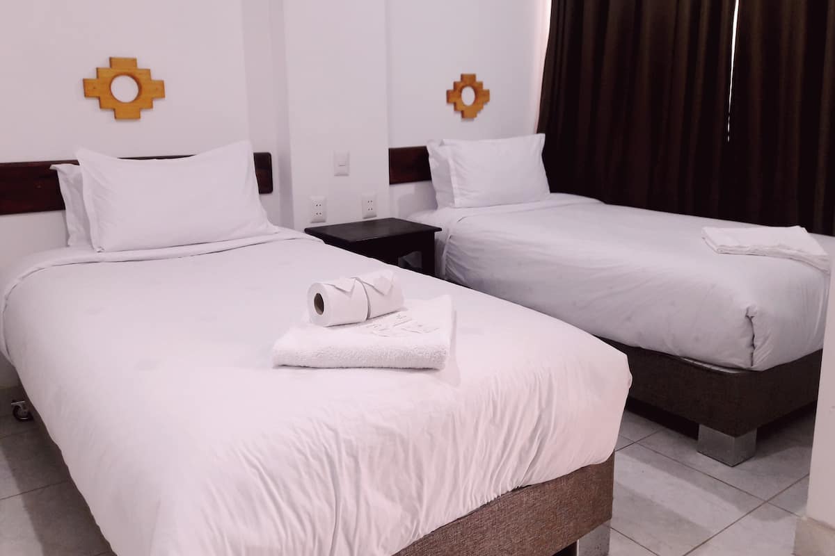 Habitació d'hotel amb dos llits, roba de llit blanca, una taula petita i una cortina.