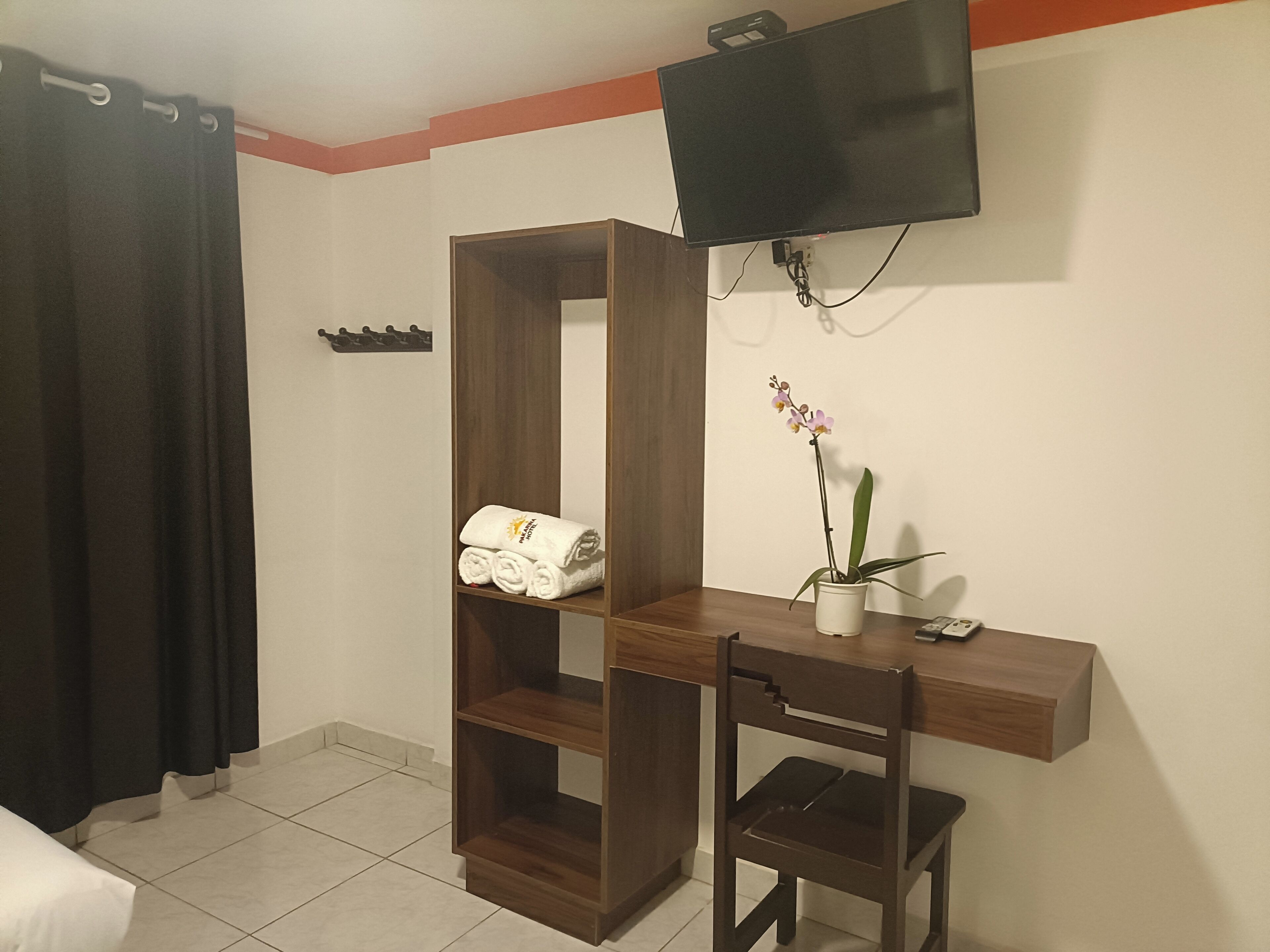 Ein Hotelzimmer mit einem Holztisch, einem Stuhl, einem an der Wand befestigten Fernseher, einer Topforchidee und einem Regal mit gerollten Handtüchern.