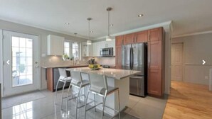A modern kitchen with stainless steel appliances, a central island with bar stools, and a view of the outdoors through a glass door. - La Maison du Bonheur 🫶 (Saint-Paul-d'Abbotsford)