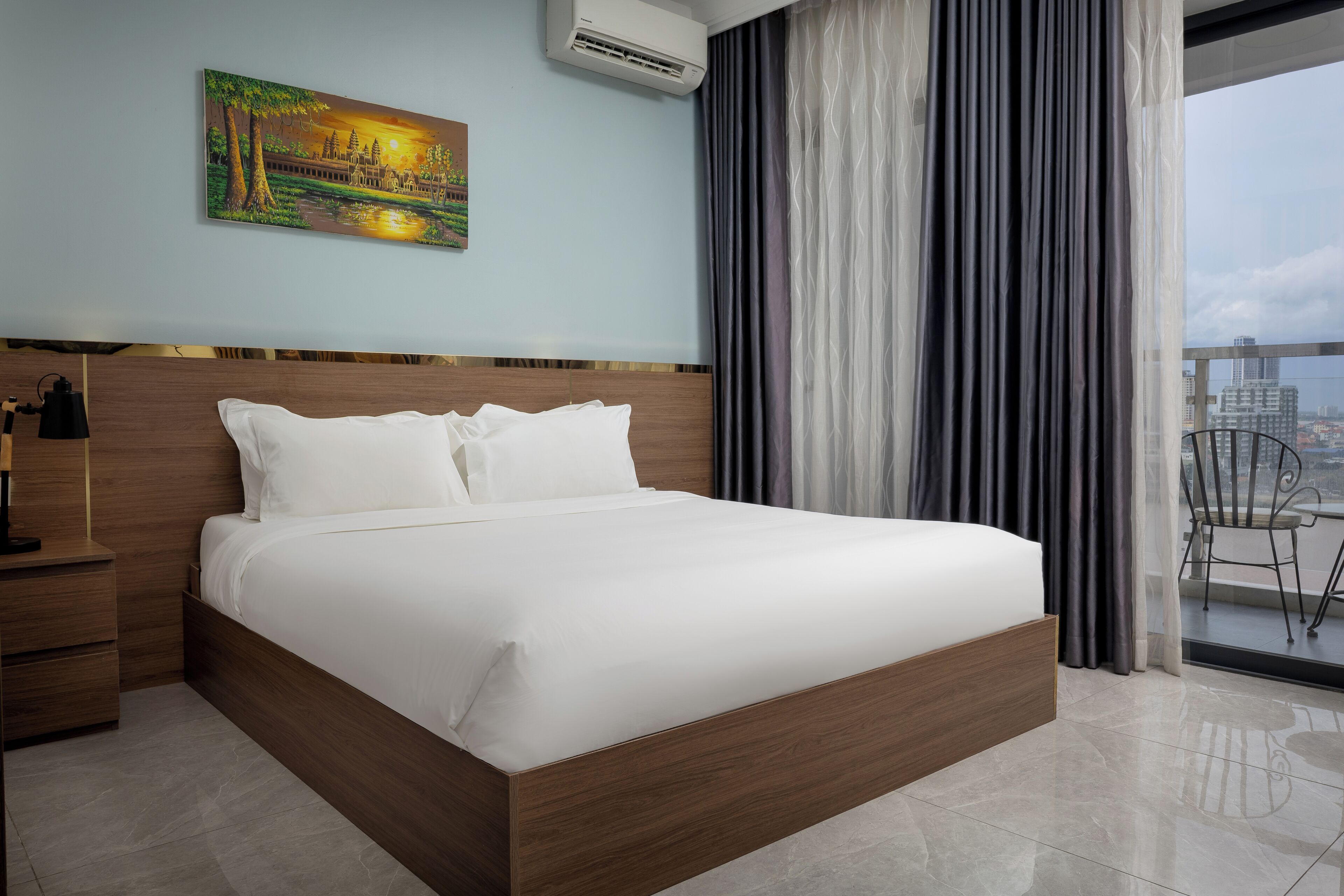 A hotel room with a large bed, a nightstand, a painting on the wall, and a view of the city through the window.