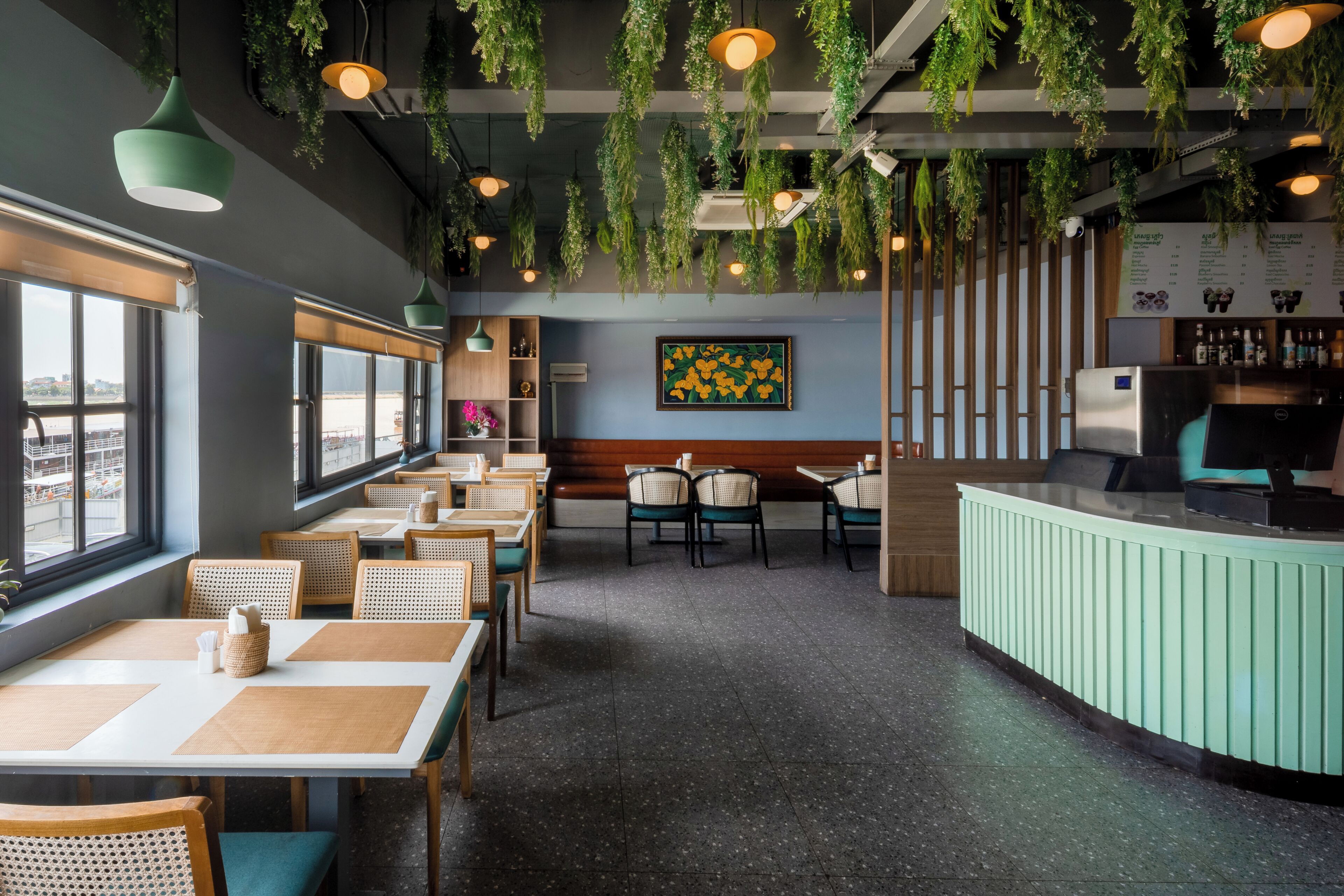 A restaurant interior with tables, chairs, hanging plants, a counter, and a menu board.