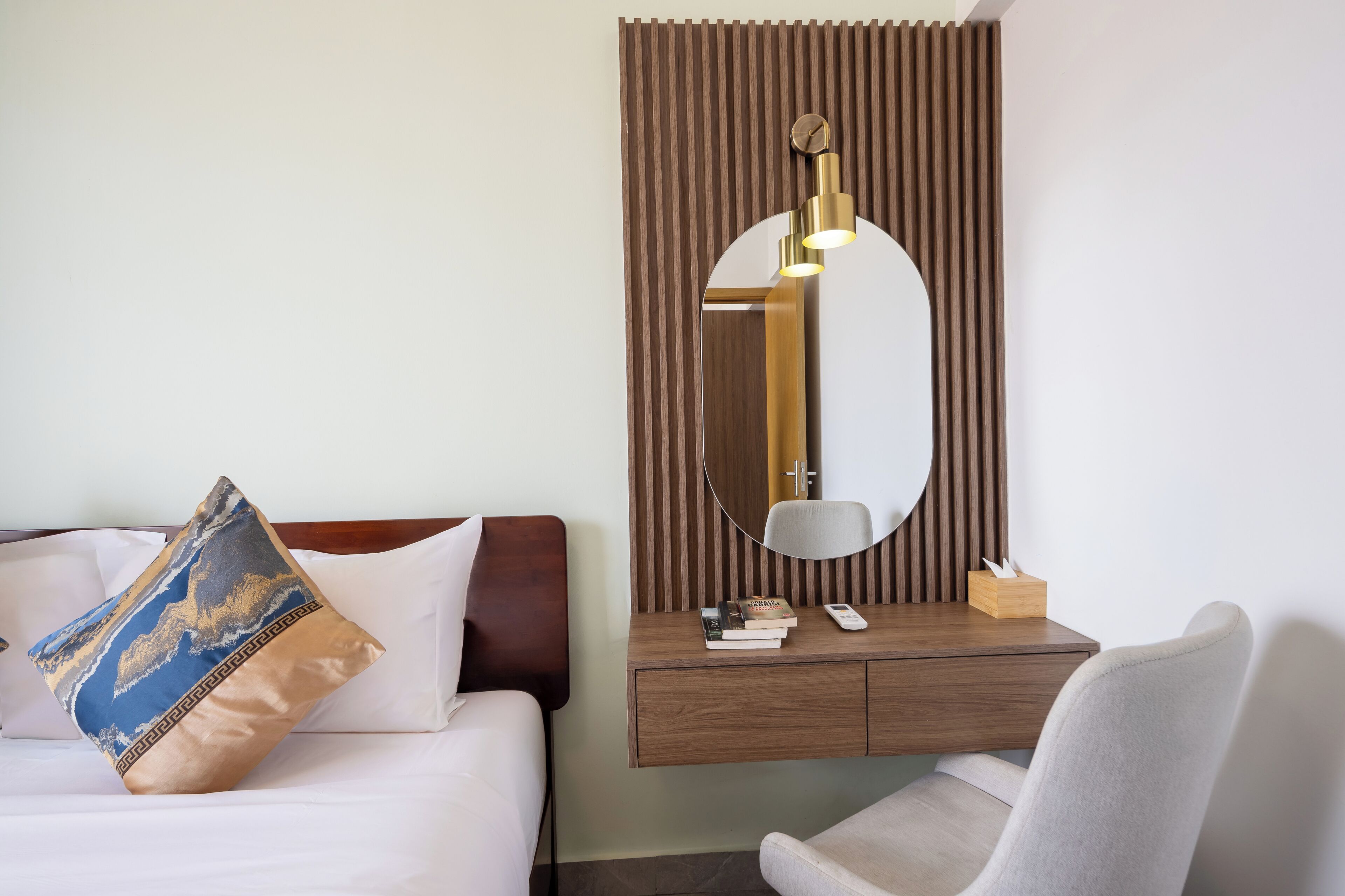 A hotel room with a bed, a bedside table with a mirror, a chair, and a small wooden box.