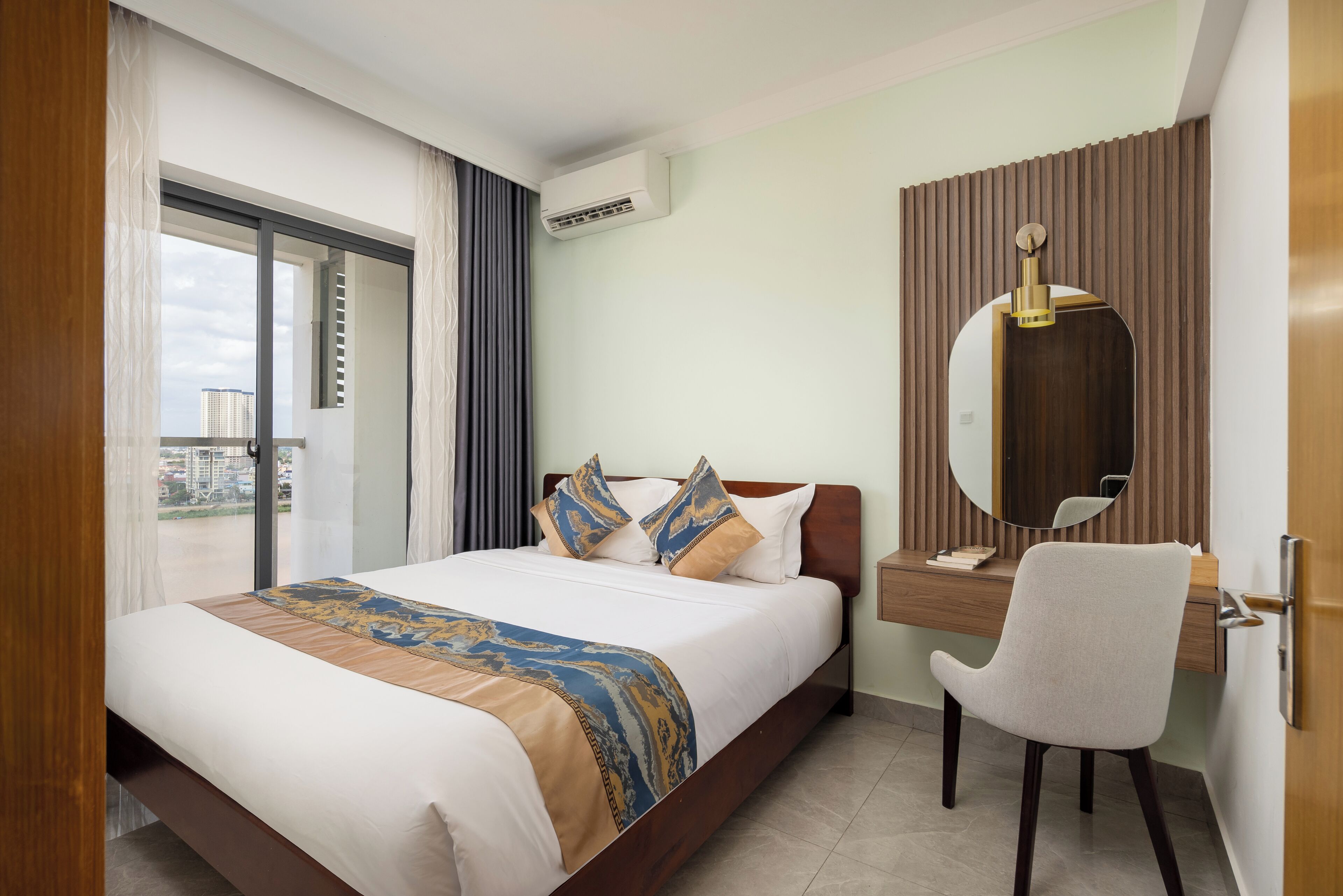 A hotel room with a bed, a desk, a chair, a mirror, and a view of the city.