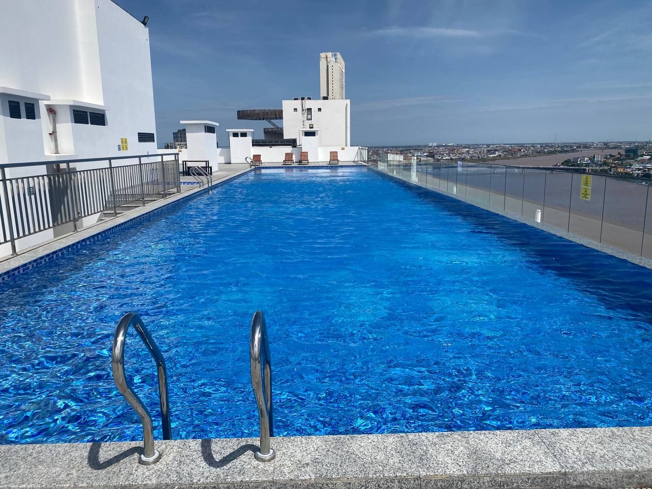 Een zwembad op het dak met helderblauw water, metalen zwemtrapjes en een uitzicht op de stadsachtergrond.