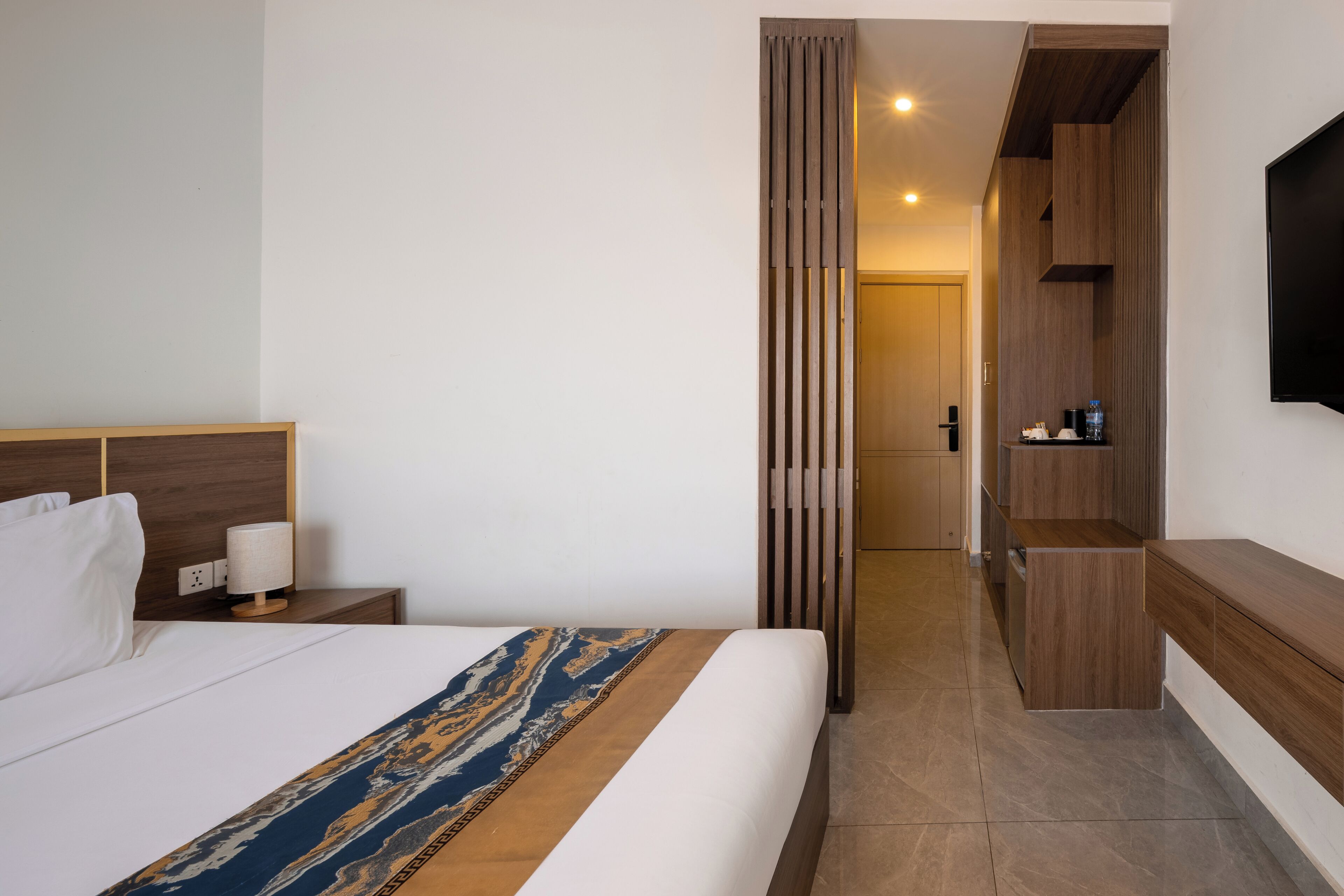 A hotel room with a bed, bedside table, lamp, and a view into the bathroom.