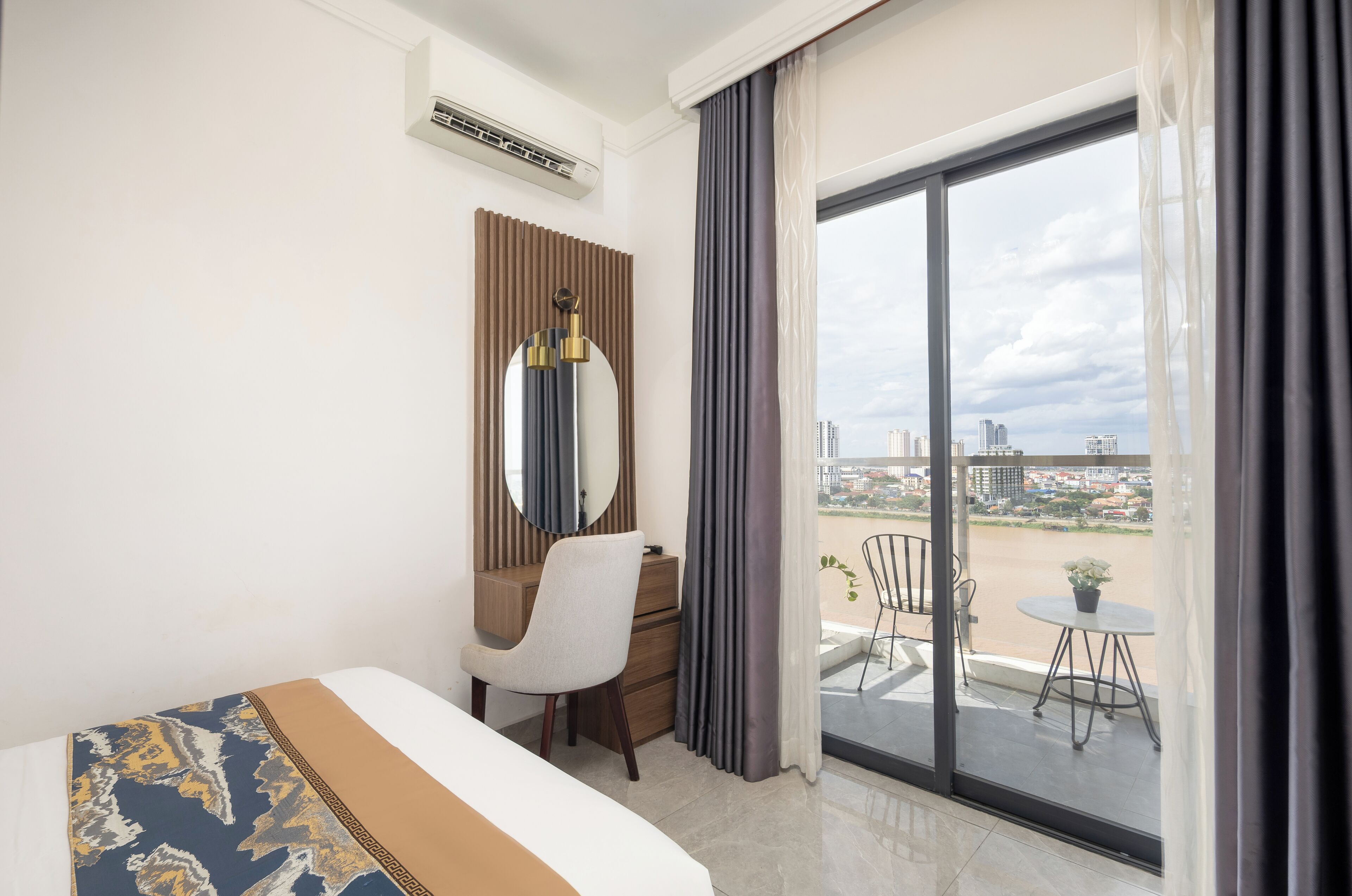 A hotel room with a bed, a desk, a chair, a large mirror, and a view of the city through a balcony.