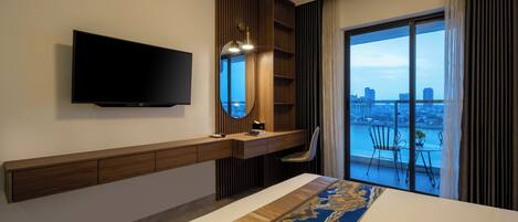 A hotel room with a bed, a TV mounted on the wall, a desk with a chair, a window with a view of the city, and a balcony with chairs.