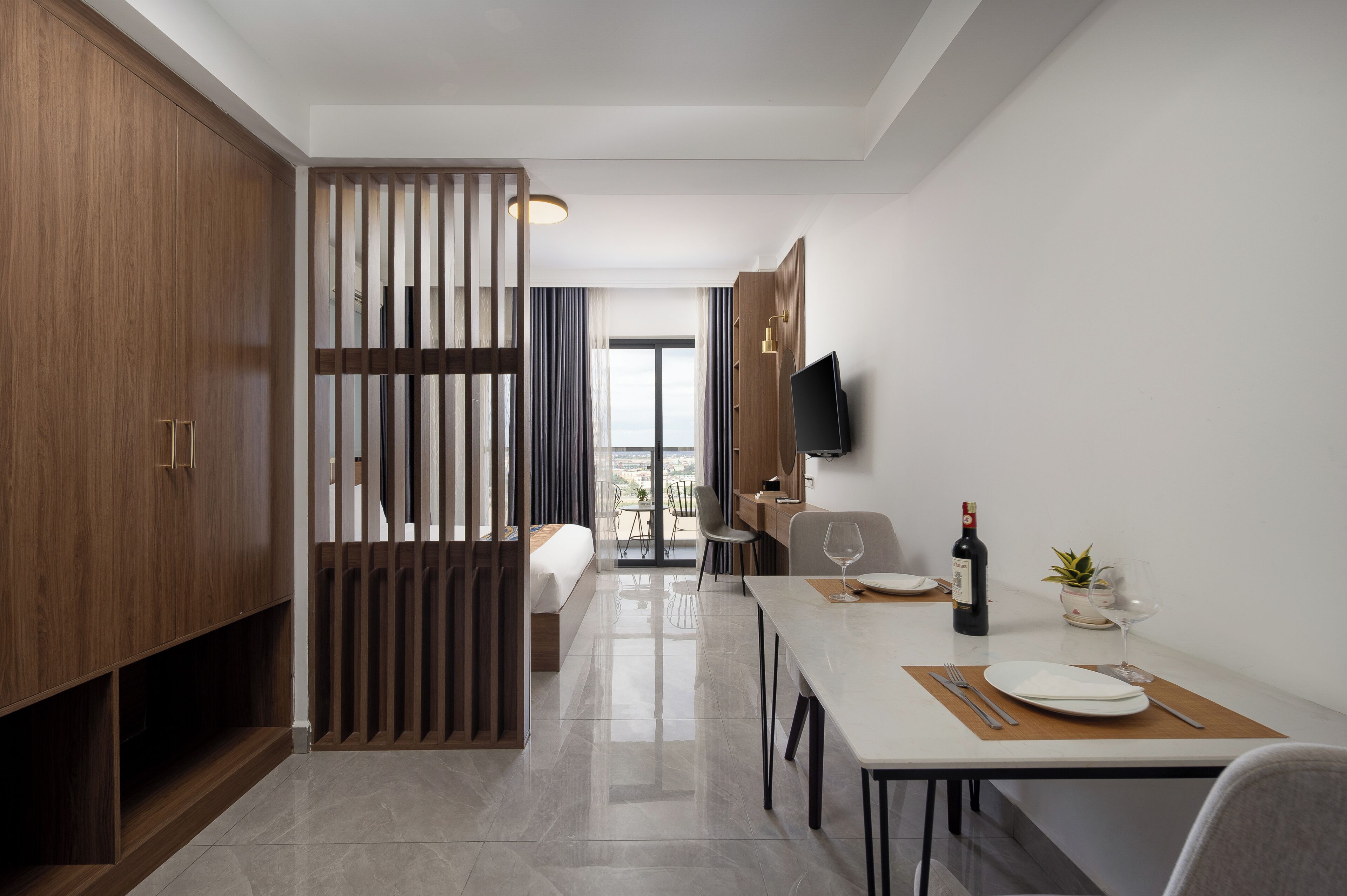 A modern hotel room with a wooden wardrobe, a dining area with a table set for two, a television mounted on the wall, and a balcony with a view.