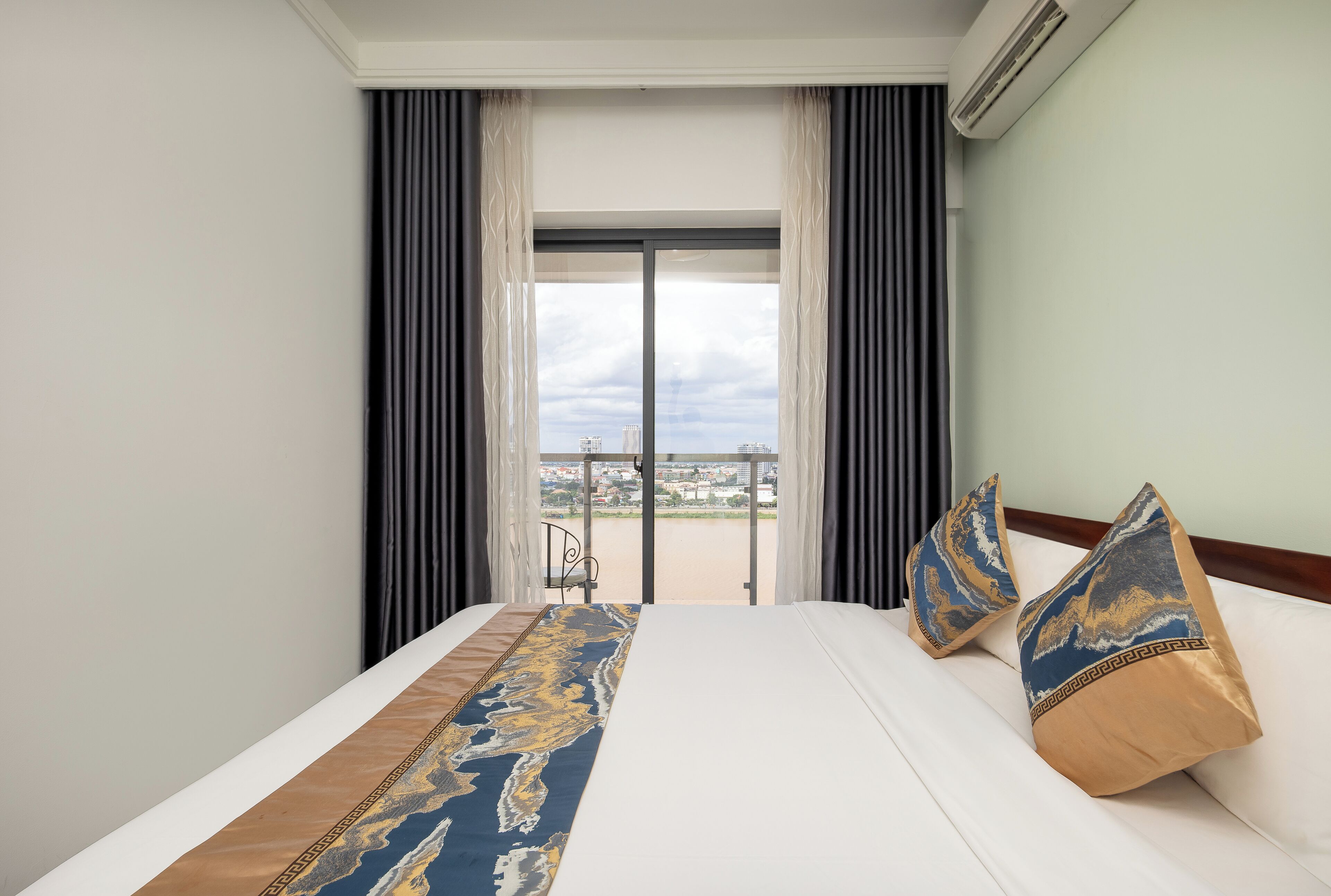 Ett hotellrum med en säng, två kuddar, utsikt över stadsbilden genom ett stort fönster och en balkong med en stol.
