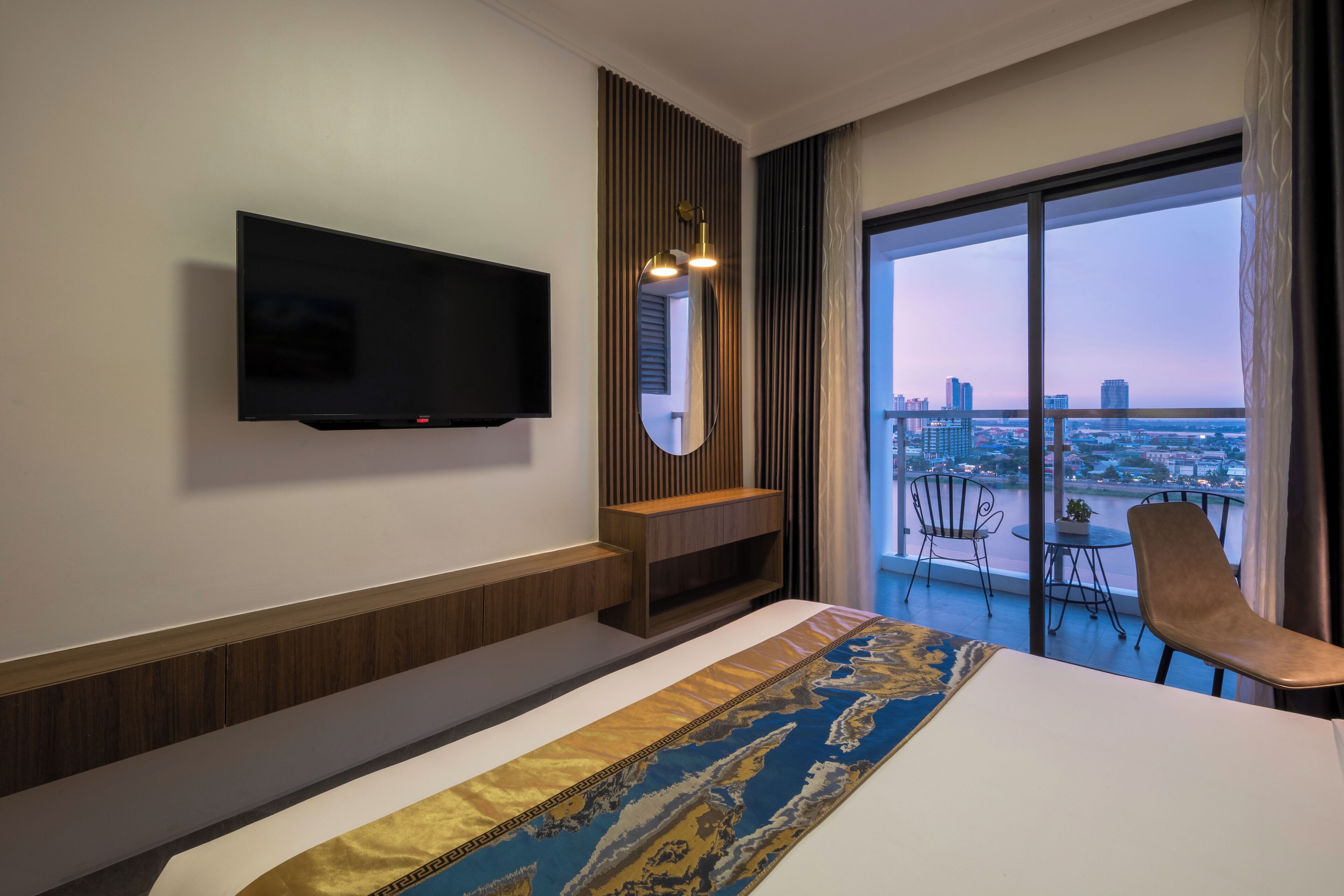 Habitación de hotel con cama, televisor montado en la pared, una pequeña mesa consola de madera, una silla y balcón con vistas a la ciudad.