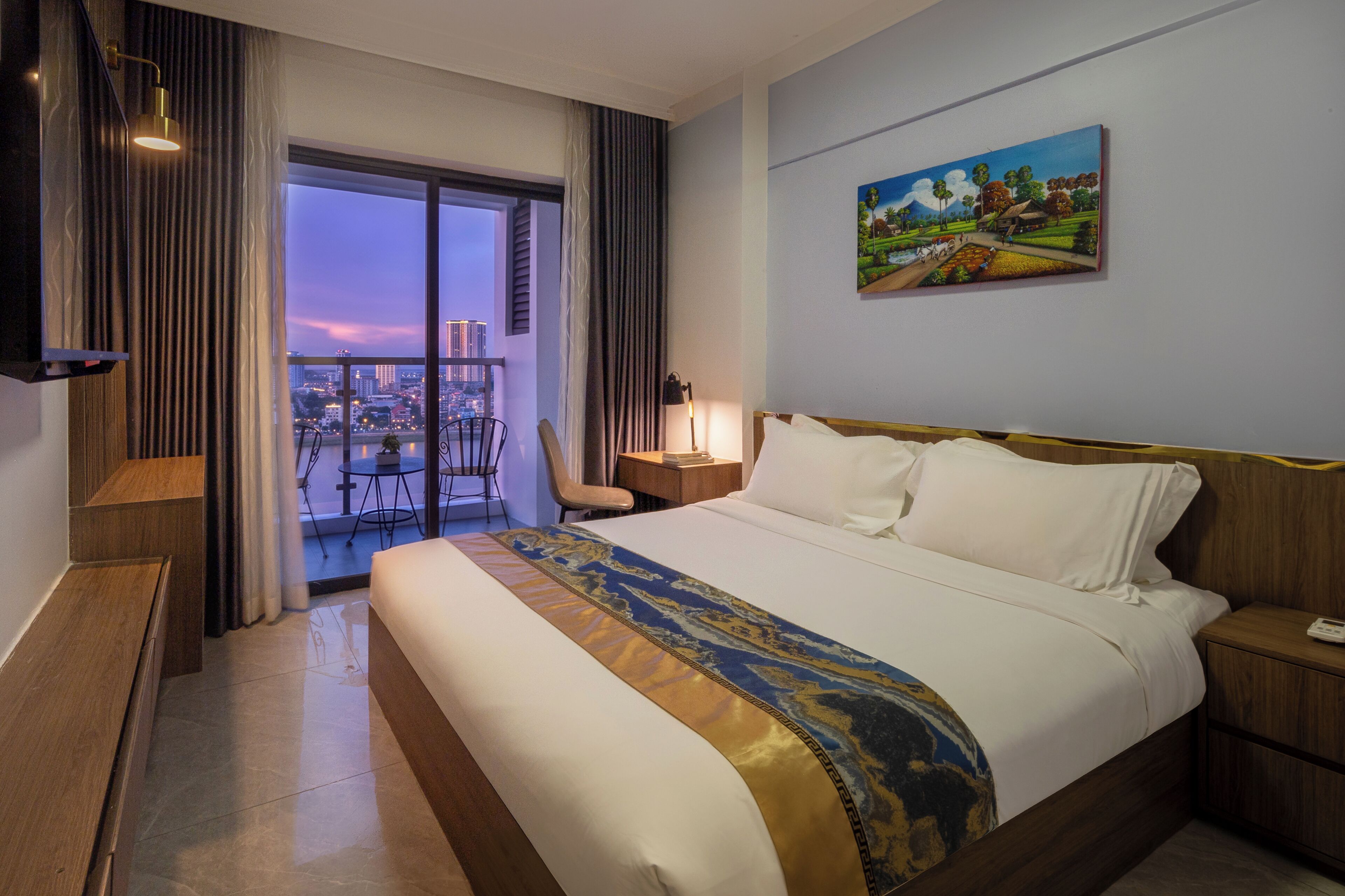 A hotel room with a large bed, a view of the cityscape through the window, a painting on the wall, and a balcony with chairs.