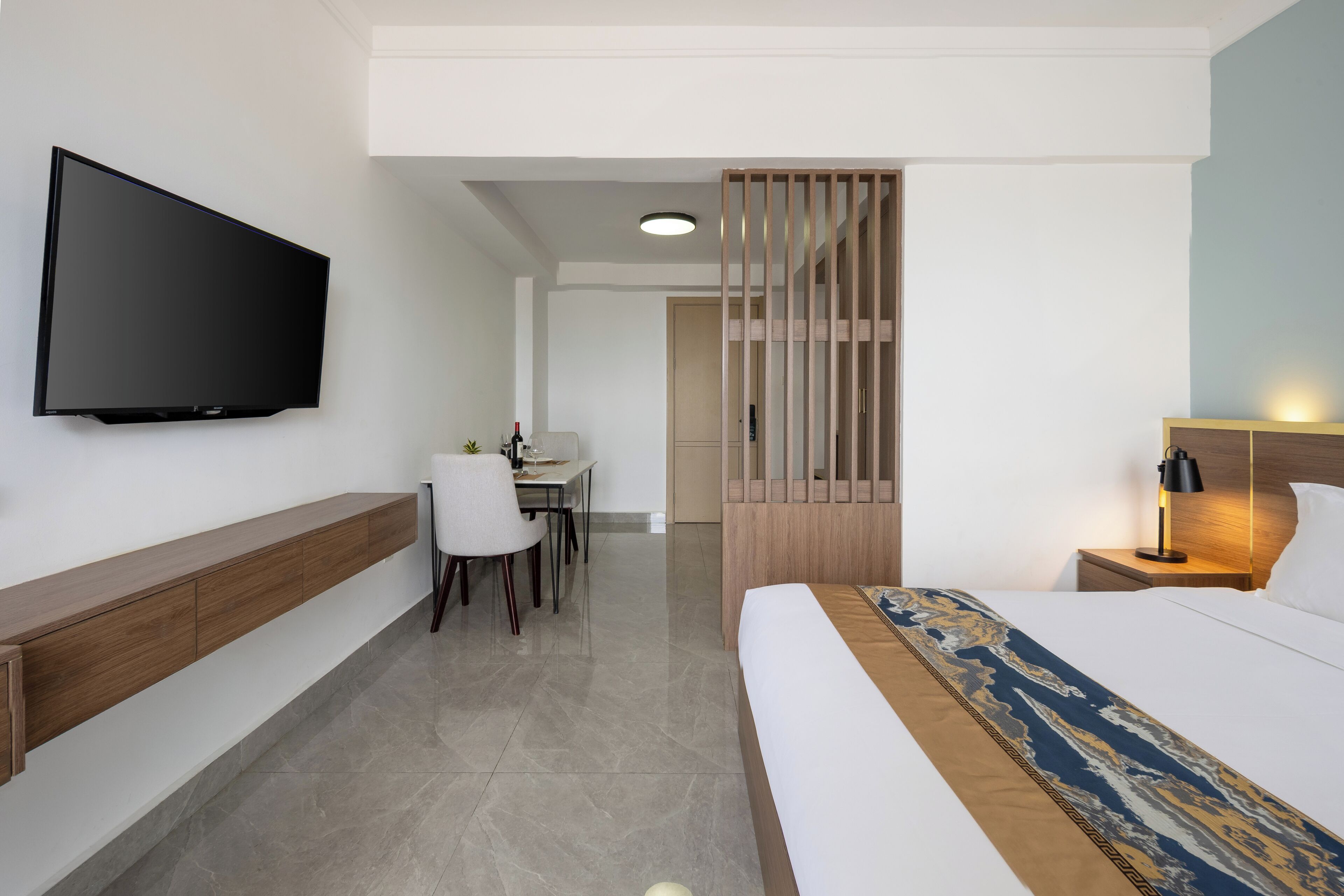 A hotel room with a bed, bedside table, TV, desk with chair, and a partition wall.