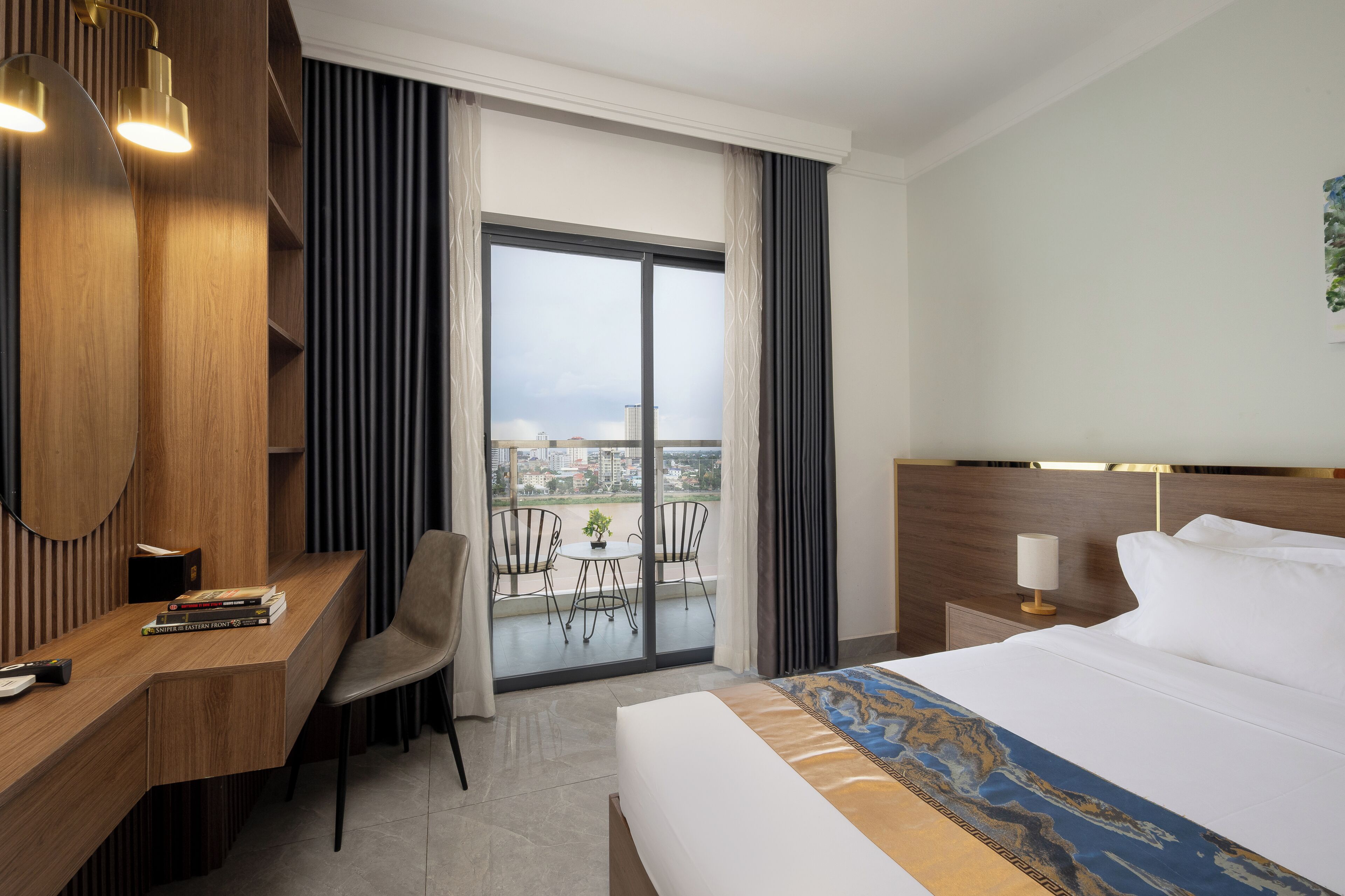 A hotel room with a bed, a desk, a chair, a balcony with a view of the city, and a window with curtains.