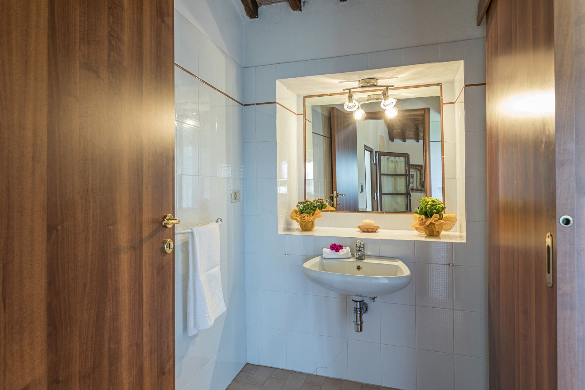 A bathroom with a wooden door, white tiled walls, a sink with a mirror above it, and two potted plants on the sink counter.