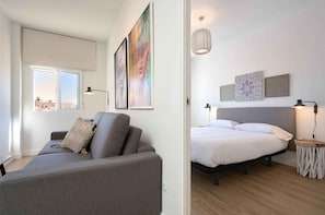 A modern living room with a sofa and a bed in the adjacent room. Both rooms have artwork on the walls and a ceiling light fixture.