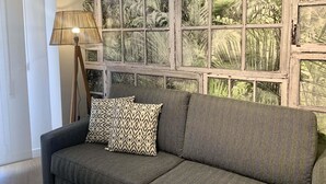 A room with a sofa, a lamp, and a large window with a tropical view. - Apartamento con Terraza y Parking Gratis (Sevilla)