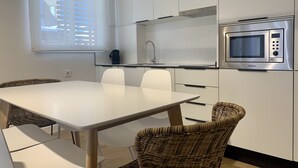 A compact kitchen with a white table, two wicker chairs, a microwave, and a window with blinds. - Apartamento con Terraza y Parking Gratis (Sevilla)