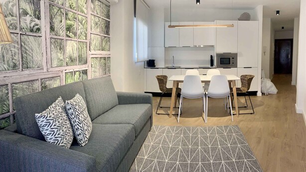 A modern living room with a grey sofa, patterned cushions, and a geometric rug. A dining area with white chairs and a wooden table is visible. A kitchen with white cabinets and a microwave is in the background. - Apartamento con Terraza y Parking Gratis (Sevilla)