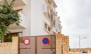 Une entrée fermée par un portail mène à un bâtiment moderne, agrémentée d’un panneau indiquant qu’il est interdit de se garer et d’un autre panneau indiquant la limitation de vitesse.