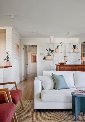 A modern living room with a white sofa, a wooden coffee table, and a red chair. There is a kitchen area with a wooden cabinet and a hanging plant.