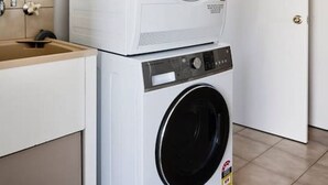 A stacked washer and dryer unit in a room with a beige tiled floor and a white door. - Peachbud House Portarlington (Portarlington)